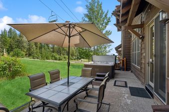 An inviting outdoor patio at Slopeside 111 features a dining table with an umbrella, a hot tub, and a view of a green lawn and trees, with a ski lift visible in the distance.