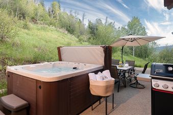 An outdoor patio at Starfall Summit featuring a hot tub, a BBQ grill, and a dining table under an umbrella with a hillside view.