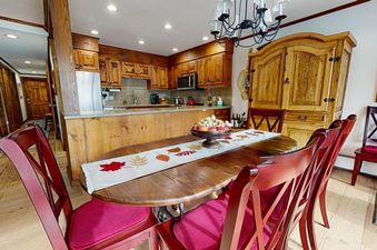 A rustic dining room features a wooden oval table with a fall-themed runner and a bowl of fruit, surrounded by red chairs, with a kitchen visible in the background.