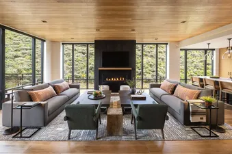 A modern and elegant living room in The Sylvan Lodge Two Bedroom Junior Penthouse with two large sofas, armchairs, a fireplace, and expansive windows showcasing a lush green hillside.