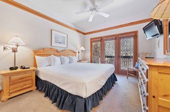 A king-sized bed with a carved wooden headboard is dressed in white linens and a deep blue bed skirt in The Charter F3305, a cozy bedroom featuring wooden furniture and access to a balcony through patterned glass doors.