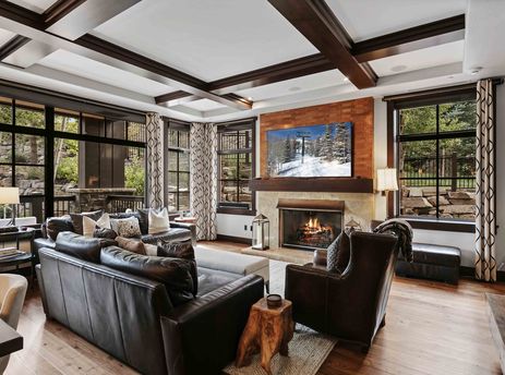 A spacious and elegantly furnished living room at Flagstaff 208, featuring dark leather sofas and armchair, a stone fireplace with a large flat-screen TV displaying a winter ski scene, and large windows with patterned curtains offering views of the outdoo