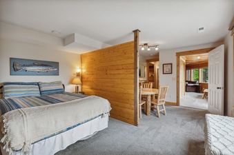 A cozy studio bedroom with a queen-sized bed, a wooden partition leading to a dining area, and an open doorway to a living room with windows.