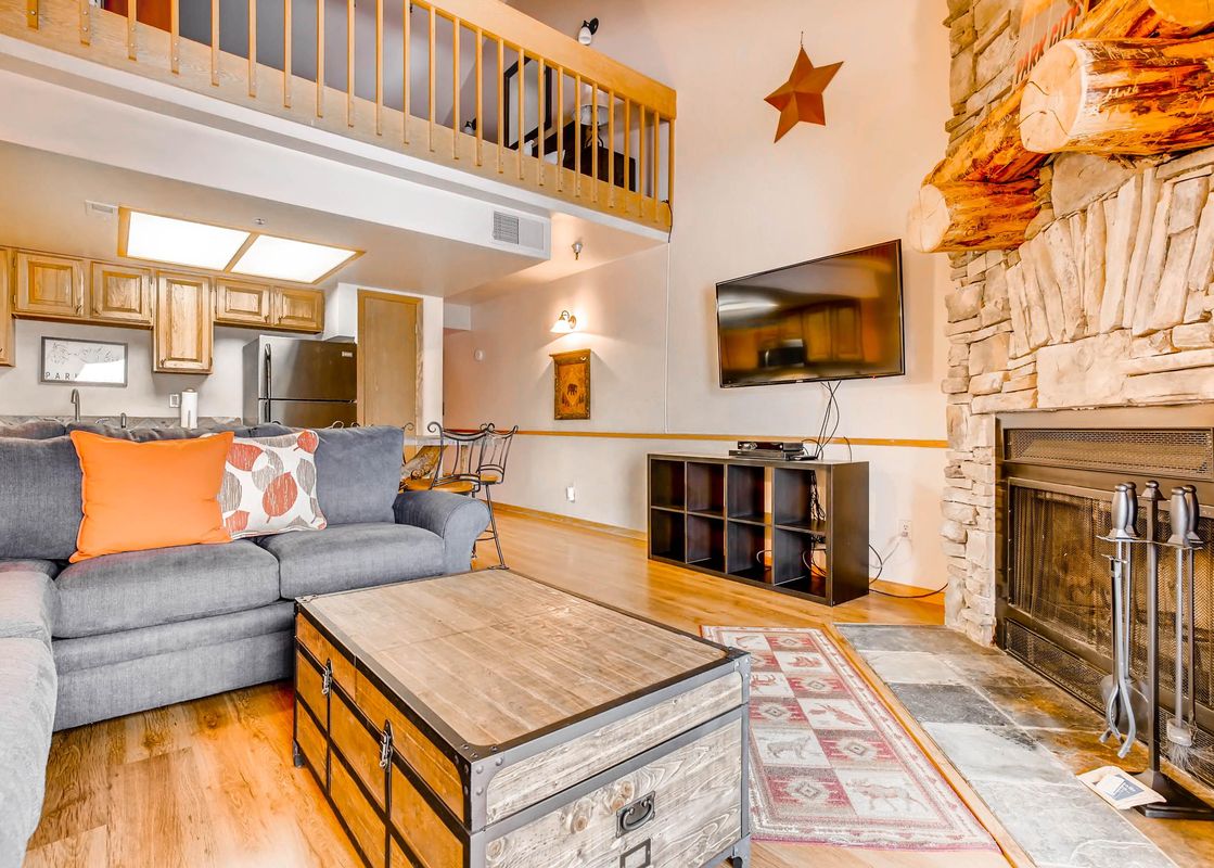 The spacious living room at Powder Pointe 303B, featuring a gray sectional, wooden coffee table, stone fireplace, flat-screen TV, and an open kitchenette with wooden cabinets.
