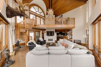 The grand living area features a plush gray sectional sofa, a spiral wooden staircase leading to a loft, a stone fireplace with a TV, and large windows, all contributing to a luxurious mountain lodge aesthetic.