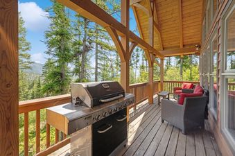 A spacious outdoor deck with a BBQ grill, wicker chairs with red cushions, and a small side table, overlooking a panoramic view of a pine forest and mountains.