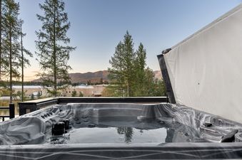 A private hot tub on a balcony offers stunning views of distant mountains and a cityscape under a soft evening sky, framed by tall pine trees.