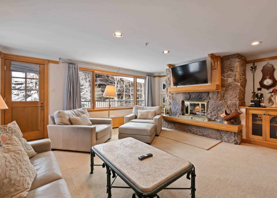 A cozy living room in Lodge Tower 387 3BR featuring a stone fireplace, a flat-screen TV, comfortable seating, and a large window with a view of a snowy winter landscape.