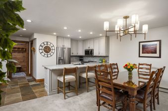 A modern kitchen with a large island and a dining area featuring a wooden table and chairs, all lit by recessed lighting and a stylish chandelier.