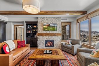 A cozy living room at Edgemont 2401 features a leather sofa, two armchairs, a stone fireplace with a TV above it, and a large window showcasing a winter mountain view.