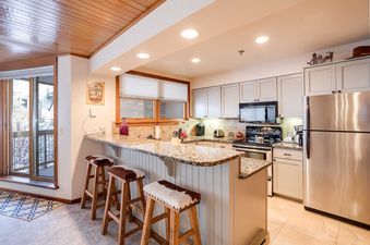 A well-equipped kitchen with modern appliances, a granite countertop, and a breakfast bar with three cowhide-covered wooden stools, illuminated by recessed lighting and featuring light grey cabinetry and a tiled backsplash.