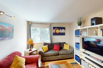 The living room of Beaver Creek Basecamp features a plush red couch with yellow pillows, a cozy gray sofa with red and yellow pillows, a coffee table with a wood top, and a large TV on the wall.