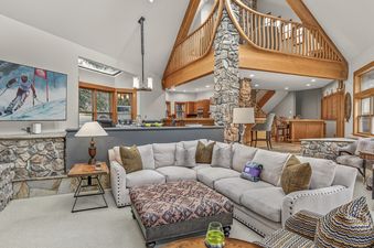 A spacious and elegantly furnished living room with a large sectional sofa, a flat-screen TV displaying a ski race, and architectural wooden beams and a stone pillar.