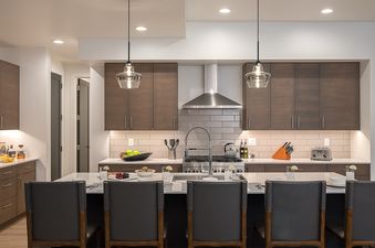 A modern kitchen at the Principal House features dark wood cabinets, light countertops, a kitchen island with gray chairs, and stainless steel appliances.