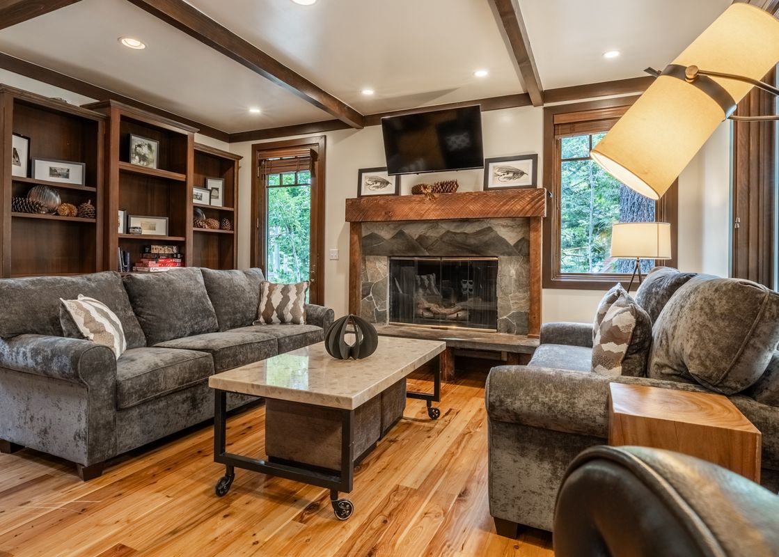 A cozy living room in Cedar Crest Owl Cottage 9 features plush gray sofas, a stone fireplace with a mounted TV, built-in bookshelves, and large windows looking out onto trees.