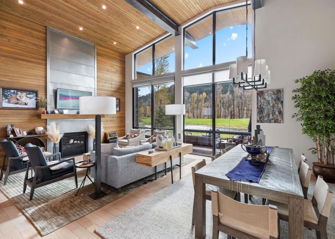 A modern cabin living and dining area with vaulted wood ceilings, a cozy fireplace, comfortable seating, and a large dining table, all overlooking a scenic golf course through expansive windows.