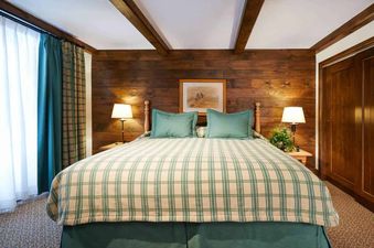 A king-sized bed with a plaid comforter and teal pillows is the focal point of a rustic bedroom with wood-paneled walls, flanked by two lamps on bedside tables, and featuring a window with curtains and a wooden closet.