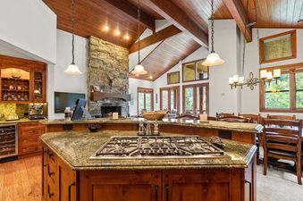 A spacious kitchen with a large island and a gas range in the foreground, leading into an open living area with a stone fireplace and large windows showcasing an exterior view.
