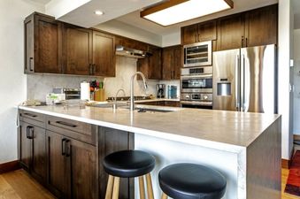 A modern kitchen with stainless steel appliances, dark wood cabinets, a granite countertop, and two bar stools.