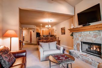 The spacious open-concept living area of Town Pointe B303 features a comfortable living room with a stone fireplace and flat-screen TV, seamlessly connected to a kitchen and dining area with wooden cabinetry and an elegant chandelier.