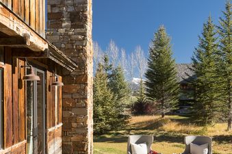An inviting outdoor stone patio at Four Pines 77 with cozy armchairs, a rustic stone chimney, and surrounding evergreens against a blue sky.