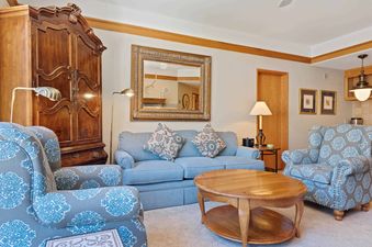 A cozy lounge at The Charter F3105 featuring a light blue sofa, two patterned armchairs, a round wooden coffee table, an antique armoire, and various lamps.