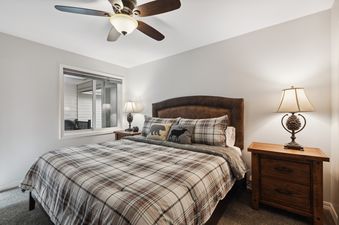 A cozy bedroom with a plaid comforter, decorative pillows, wooden nightstands, and lamps, along with a ceiling fan and a window.