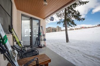A private patio with skis leaning against the wall, outdoor seating, and a snowy slope with buildings in the background, a gondola, and a blue sky.