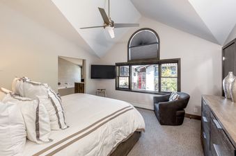 The bedroom at Vail Center Place 3 Bedroom Diamond Unit 3 features a large bed with white and gray bedding, a flat-screen TV, a cozy armchair by the window, and a ceiling fan.