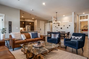 The living area within Hideaway Haven features a large leather sectional, a glass coffee table, and two blue armchairs, with a view of the kitchen and dining area in the background.