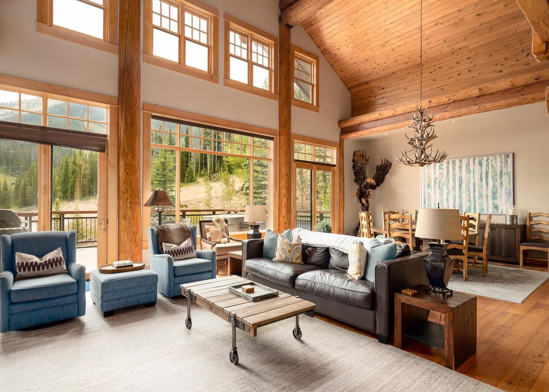 Moonlight Basin Penthouse 3 living room with large windows overlooking the forest, a leather couch, two armchairs, a coffee table, and a wood-burning fireplace.