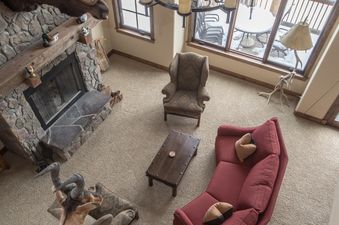 A cozy, rustic living room at 60 Moose Ridge features a large stone fireplace, plush seating, and large windows with a view of snow outside.