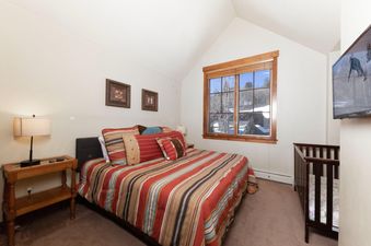 The bedroom at Villa at Mountain Thunder features a comfortable bed with a striped comforter, a bedside table with a lamp, a crib, and a large window offering a view of a snowy landscape.