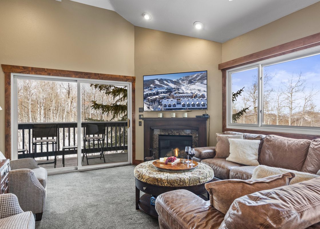 A living room with a leather sectional sofa, a coffee table, and a fireplace. The room has large windows with views of the mountains and trees.