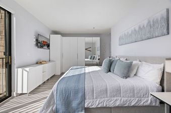 A bright bedroom at The Mason 2003 2005 with a large bed, white bedding, and blue throw, featuring a mounted TV, white storage console, mirrored wardrobe, and a balcony door.