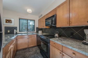 A kitchen with stainless steel appliances, wood cabinets, granite countertops, and a window with a view of trees.