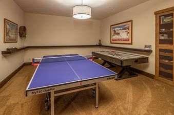 A spacious game room at The Last Best Chalet features a blue ping pong table, an air hockey table, and rustic wooden shelving with decorative items, all under warm ceiling lighting.