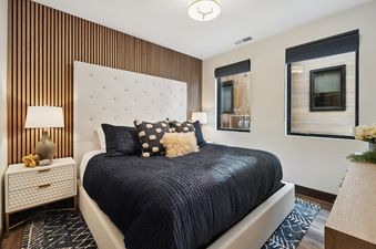 The primary bedroom features a white upholstered bed with a black duvet, flanked by a modern nightstand with a lamp and a unique wooden slat wall.