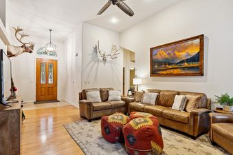 A spacious living room at Meadow Lake Villa with light wood flooring, a large area rug, two brown leather sofas with decorative pillows, two patterned ottomans, a ceiling fan, and rustic wall decor including a deer head mount and a large landscape paintin