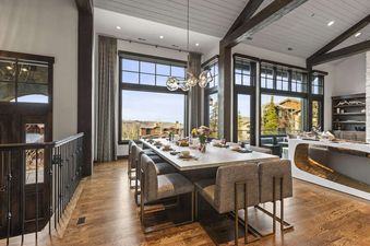 A modern dining room at Ruby Run features a large rectangular table set for a meal, surrounded by upholstered chairs with gold frames, and large windows offering a scenic view of a mountainous landscape.