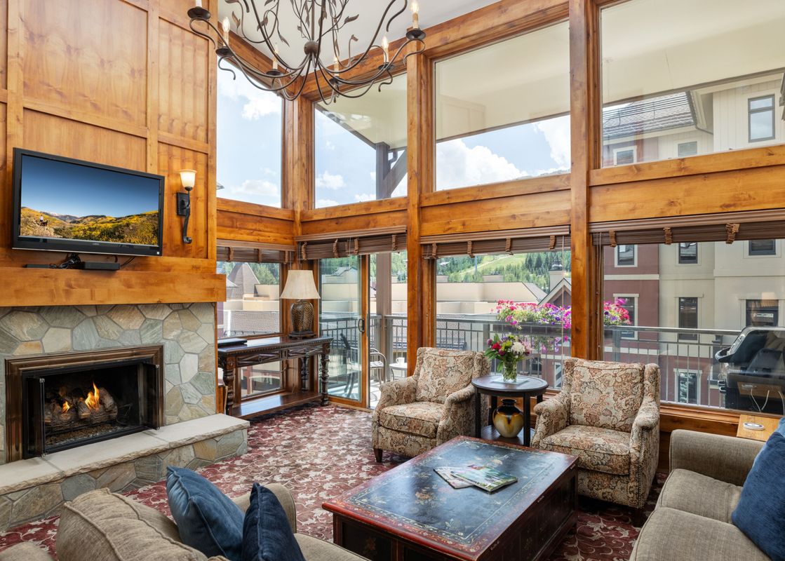 A cozy living room in Montaneros 418 features a stone fireplace, a wall-mounted TV, and comfortable seating with large windows looking out to the surrounding buildings and nature.