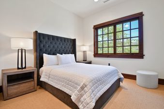 A cozy bedroom with a dark tufted bed, white bedding, wooden nightstands with lamps, and a large window overlooking a lush outdoor landscape.