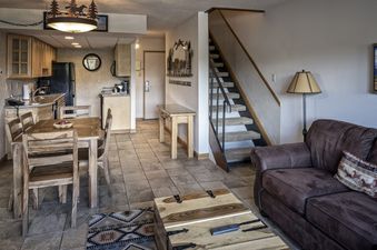 A spacious and inviting open-plan living area at the Two Bedroom Suite Beaver Run Resort, featuring a well-equipped kitchen, a wooden dining table, a comfortable leather sofa, and a rustic coffee table, all under warm lighting.