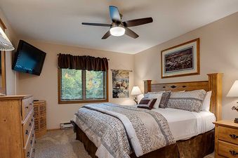 A serene bedroom at Ranch at Steamboat RA401 features a comfortable bed, rustic wooden furniture, a ceiling fan, and a wall-mounted TV, with a window offering a view of the outdoors.