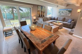 A spacious living area inside Vail International 002 features a large sectional sofa, a coffee table, and a dining area with a wooden table and several chairs, all bathed in natural light from expansive windows.