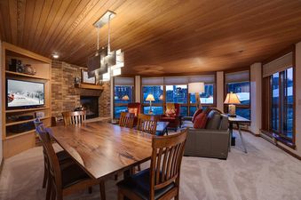 A spacious dining area with a large wooden table and chairs, leading into a cozy living room with a fireplace and large windows showcasing an exterior view at Chateau Chamonix CX123.
