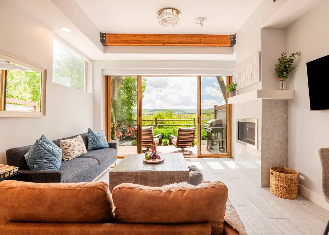 A spacious and modern living room featuring a comfortable sofa and armchairs, a large coffee table, and a fireplace, with a view of lush greenery and mountains through large glass doors.