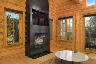 A cozy living area at Sundance Lodge 108 Lodges Lane with a modern black fireplace, log walls, large windows showcasing green outdoors, and a round white coffee table.