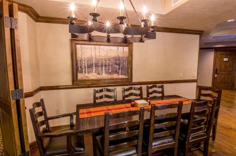 A formal dining room features a large dark wooden table with an orange and red checkered runner, surrounded by ten dark wooden chairs with cushioned seats.