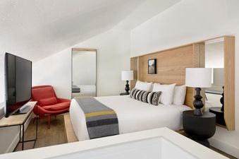 A modern lofted bedroom in the Gravity Haus JH Deluxe Loft featuring a king-size bed, sleek wood paneling, a red accent chair, and a flat-screen TV under a sloped ceiling.